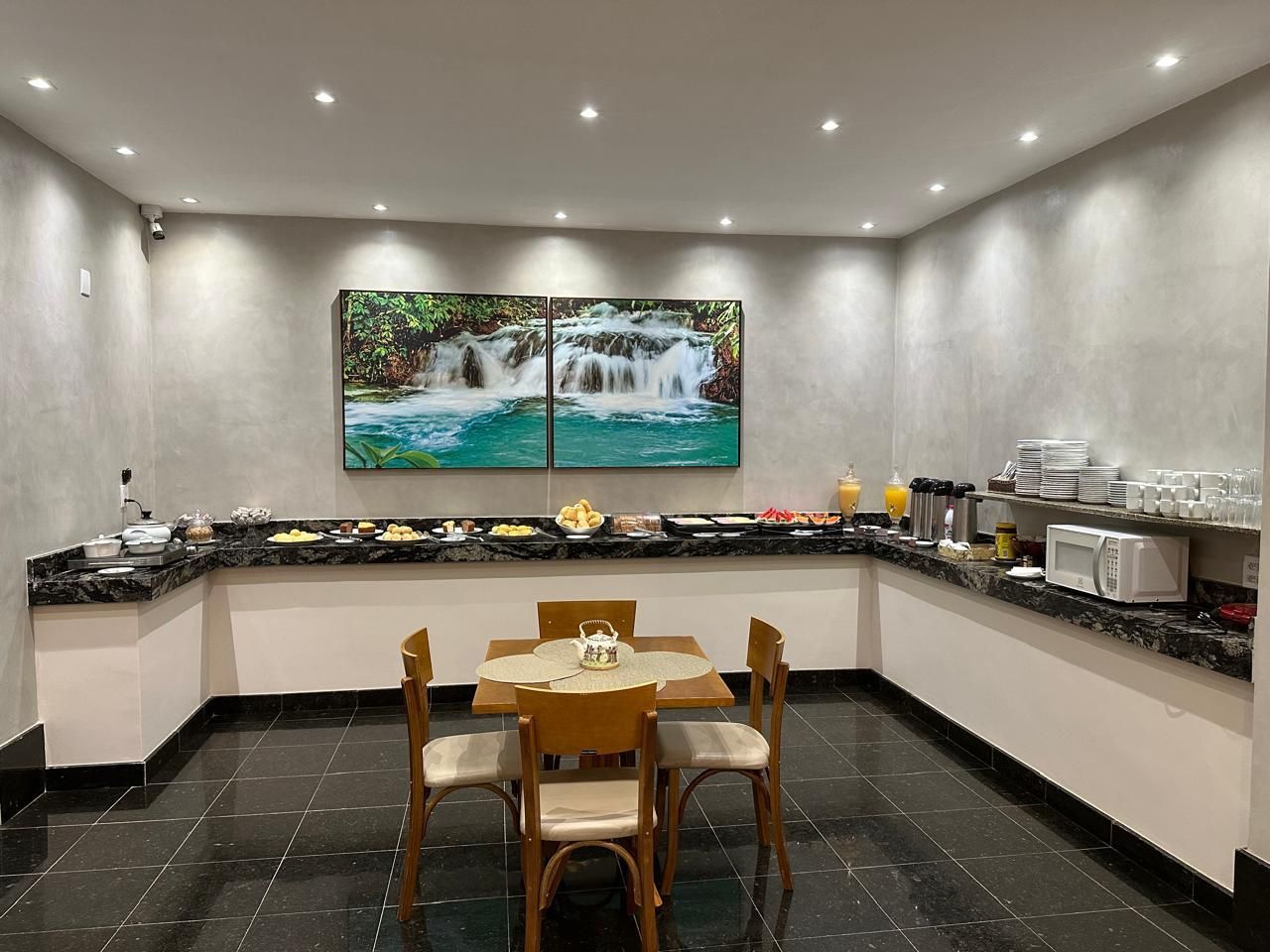 Área de buffet de café da manhã com exposição de alimentos em balcão e mesa pequena com cadeiras.