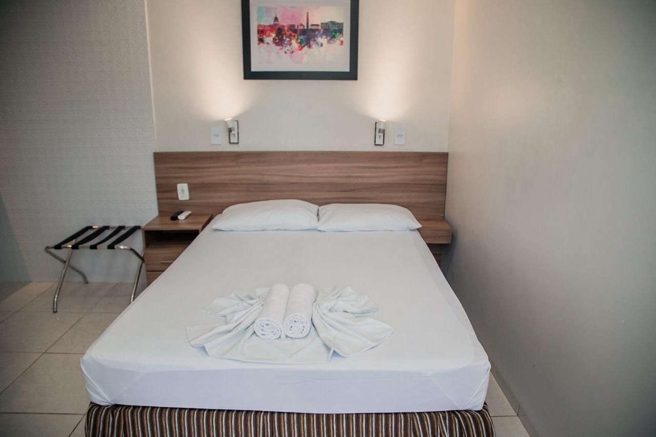 Quarto de hotel com cama, mesa de cabeceira, suporte para malas, obras de arte e arandelas.