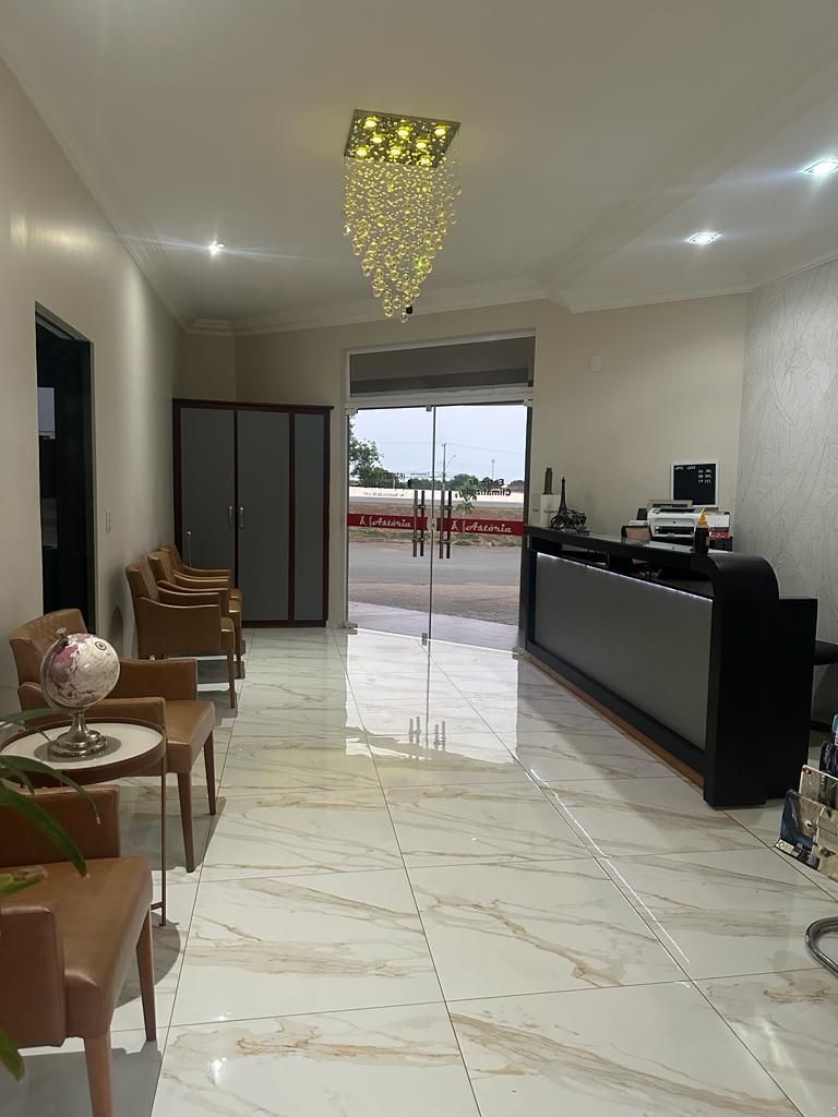 Sala de espera com cadeiras bege, armário cinza e balcão de recepção com lustre. Vista externa através da porta de vidro.