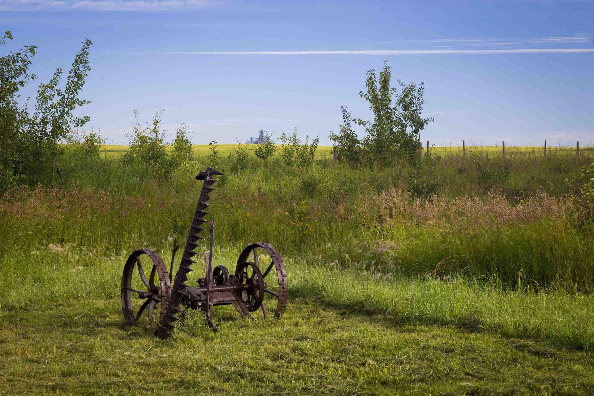 Old rusty farming scythe on a green field, with yellow and green fields and blue sky in the background.