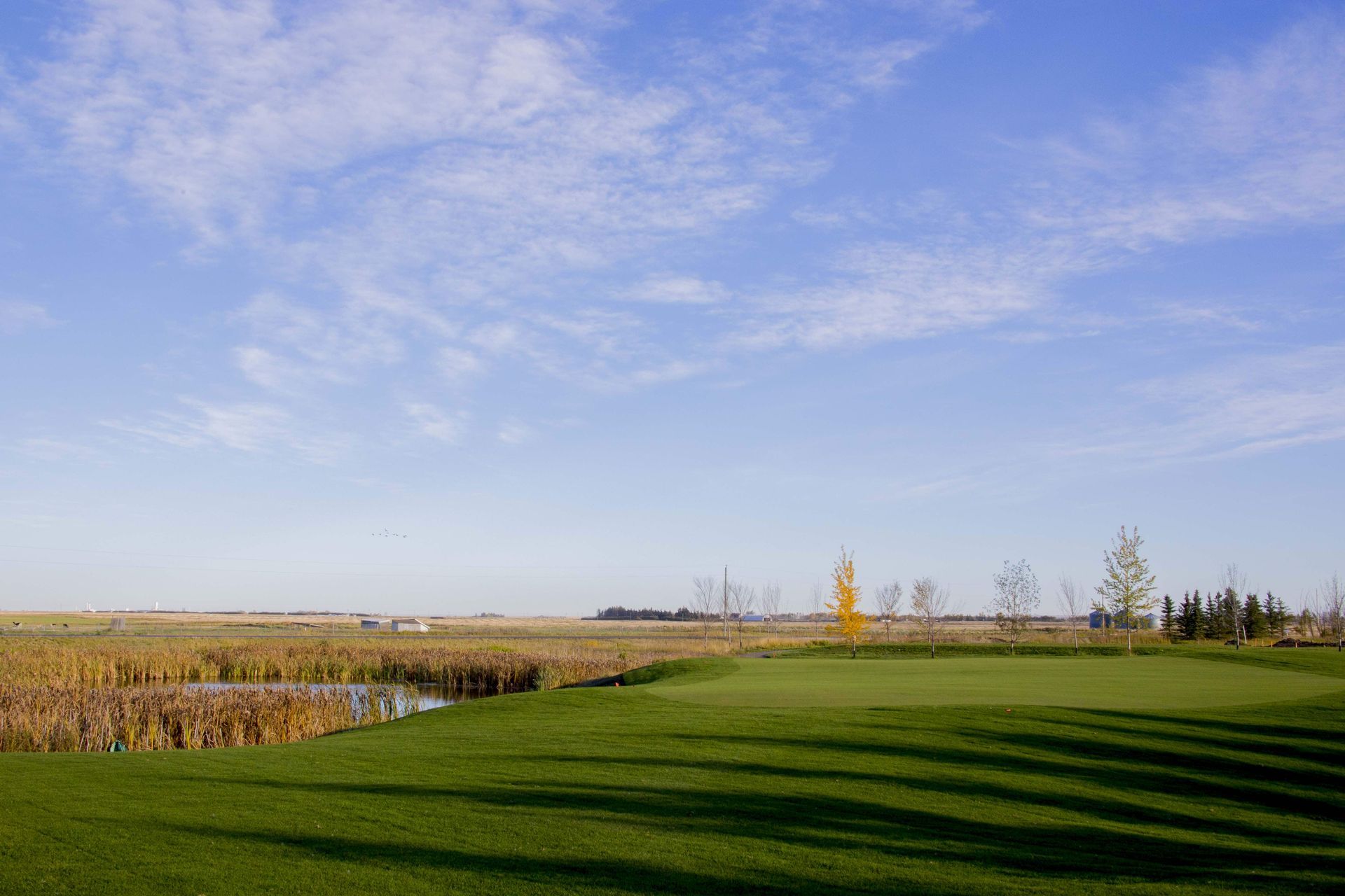 Green golf course with a pond on the left, under a blue sky with scattered clouds.