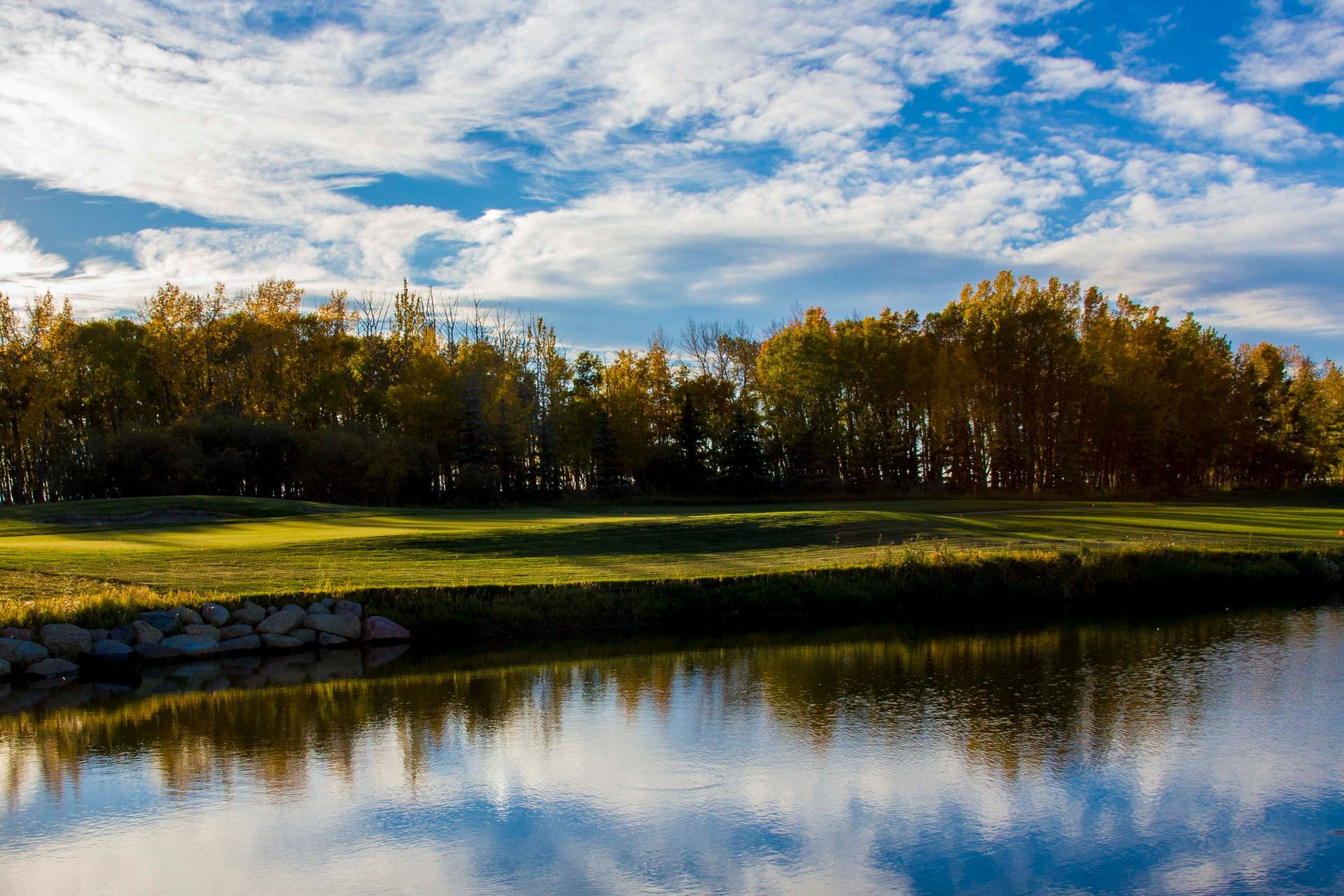 Lush green field with golden trees, reflected in calm water under a blue sky with clouds.