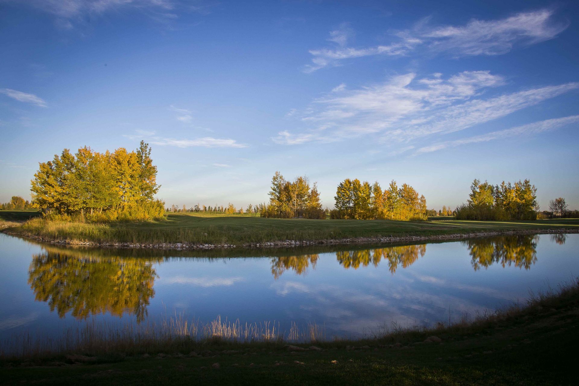 Calm water reflects trees with yellow leaves under a blue sky with clouds.