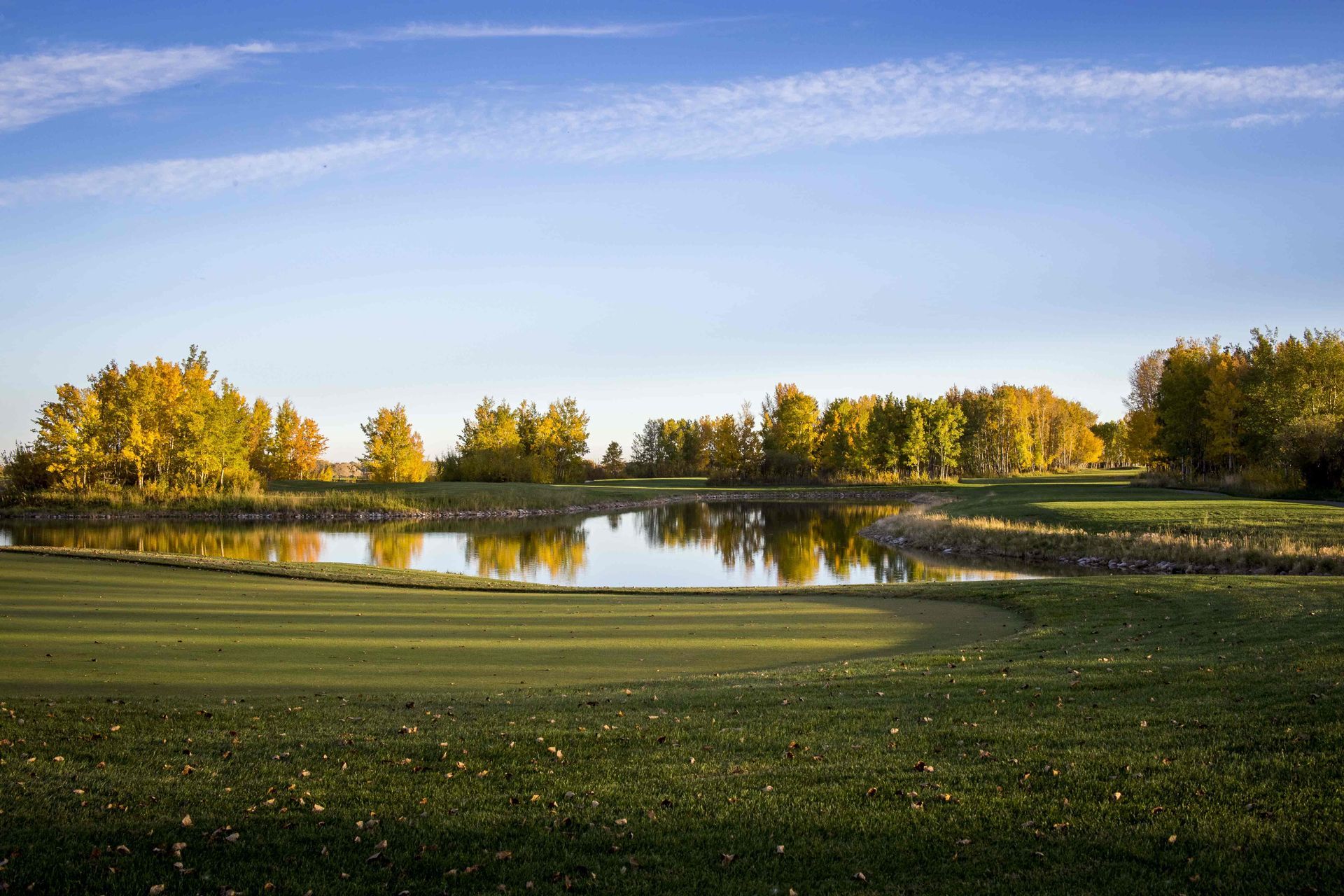 Green golf course with a pond, surrounded by trees with yellow autumn foliage under a blue sky.