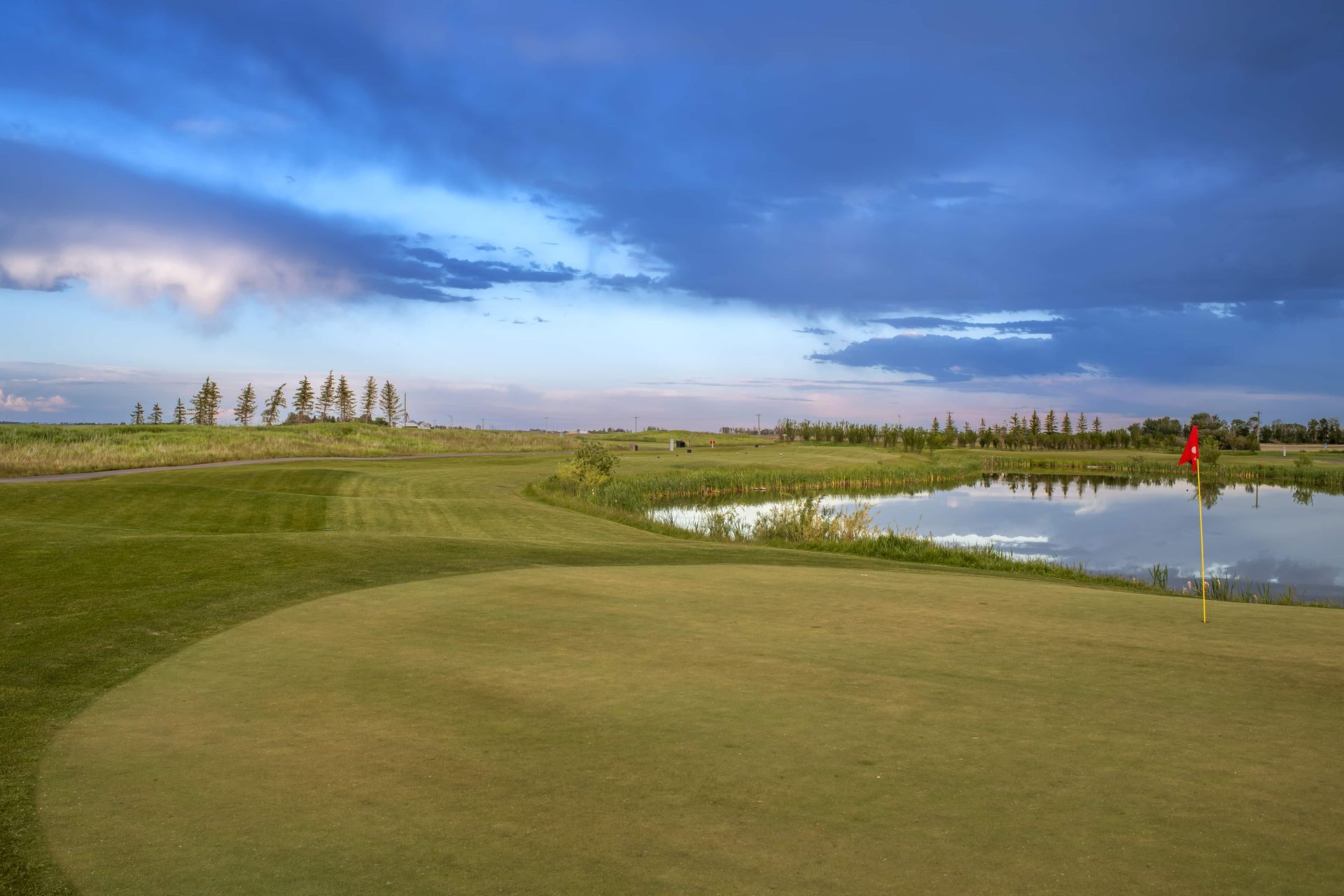 Golf green beside a pond under a cloudy sky. Red flag marks the hole.