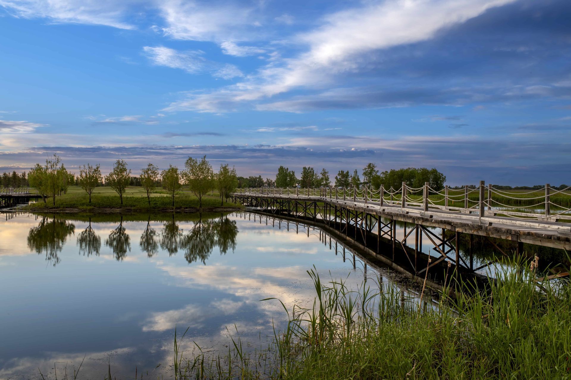 A wooden bridge curves over a still lake, reflecting trees and a cloudy sky at sunset.