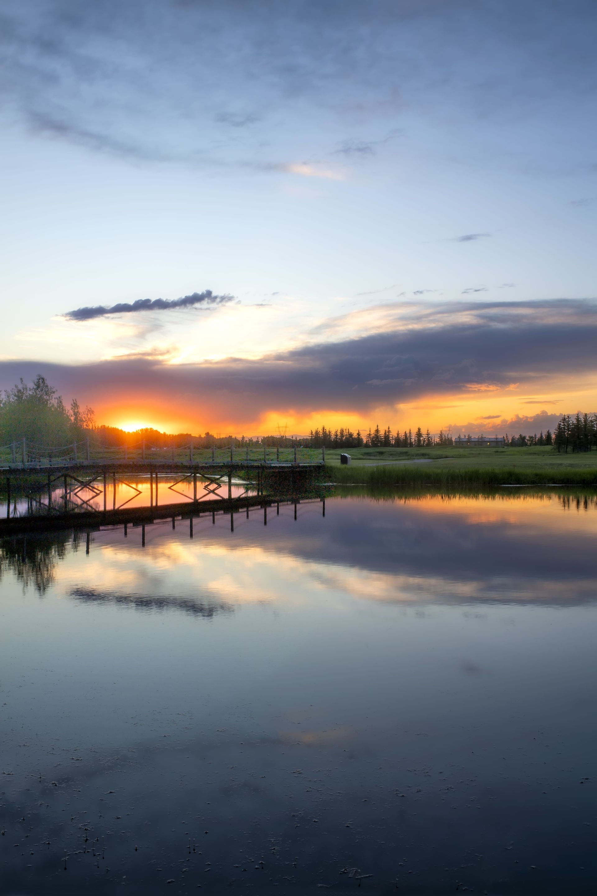 Sunset over a calm lake reflecting the sky and a wooden bridge.