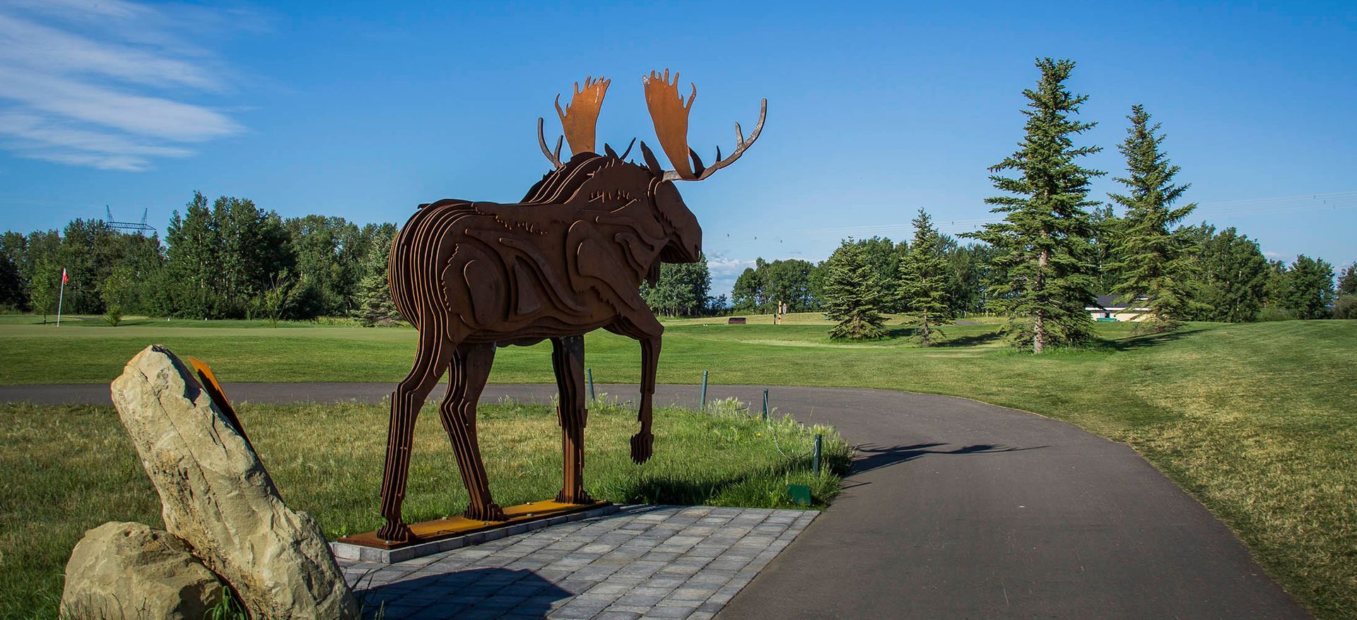 Metal moose sculpture on a path in a park with trees and blue sky.