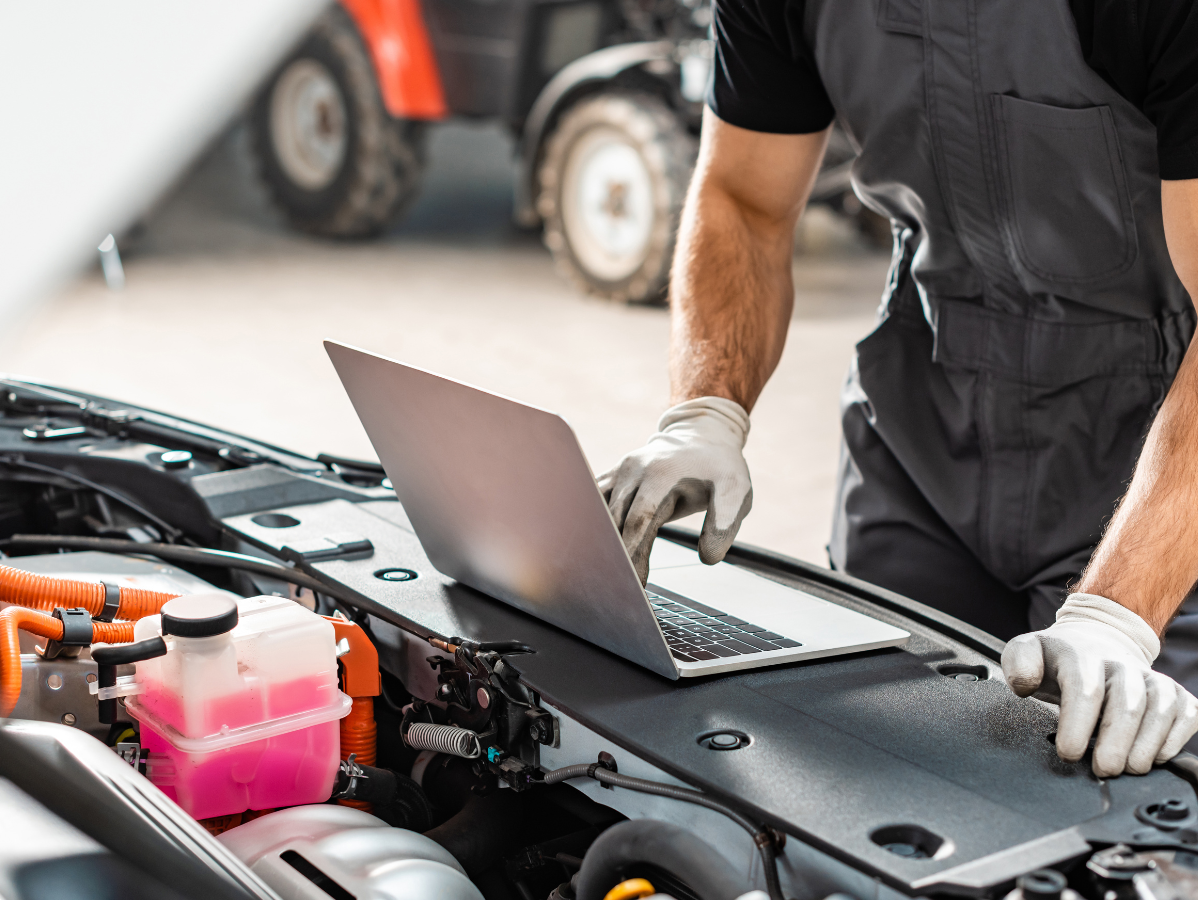 Mechanic using laptop to diagnose a car engine with hood open, wearing gloves, in a garage.