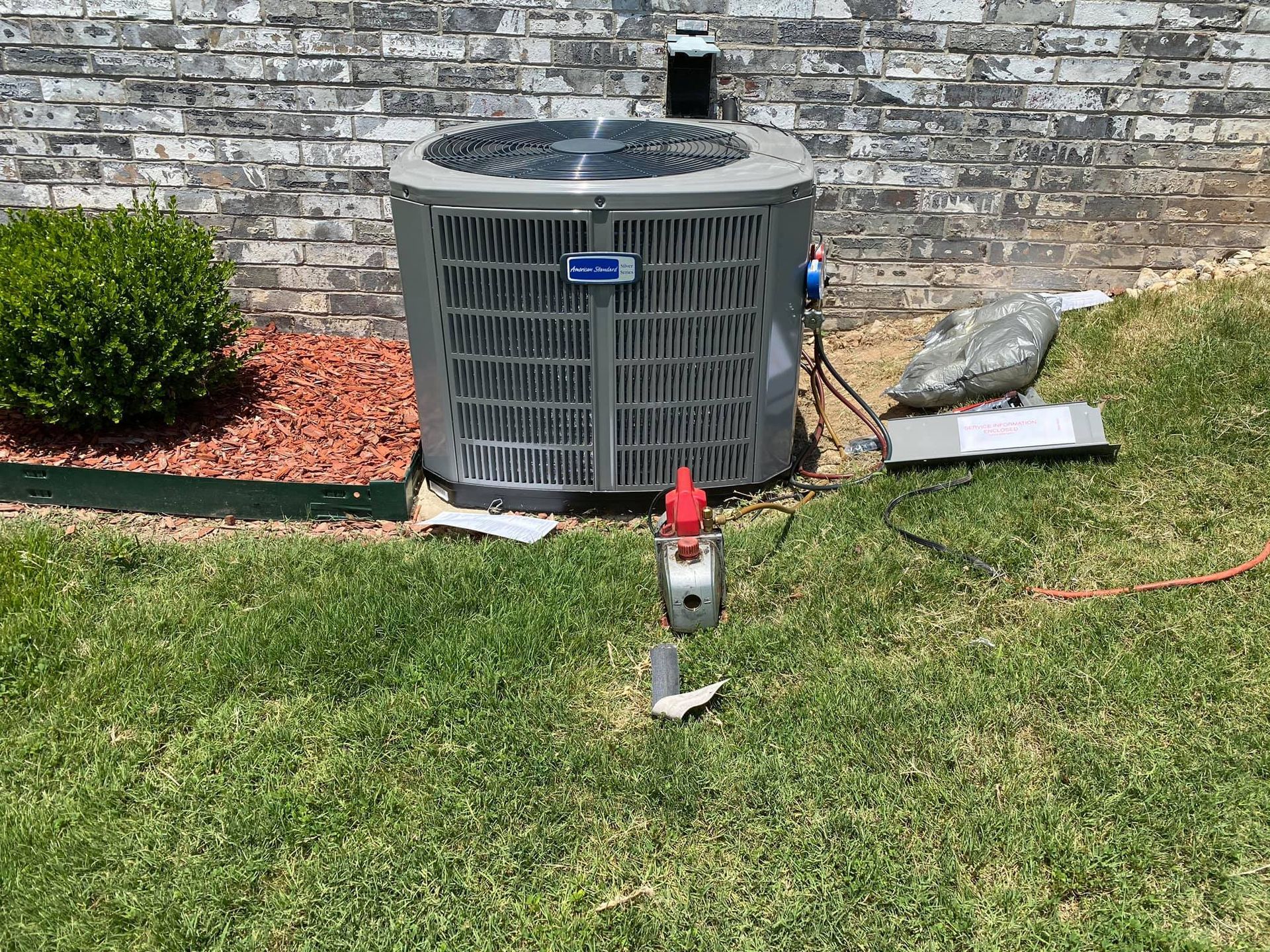 An air conditioning unit sits on grass next to a brick wall, with red and gray components visible.