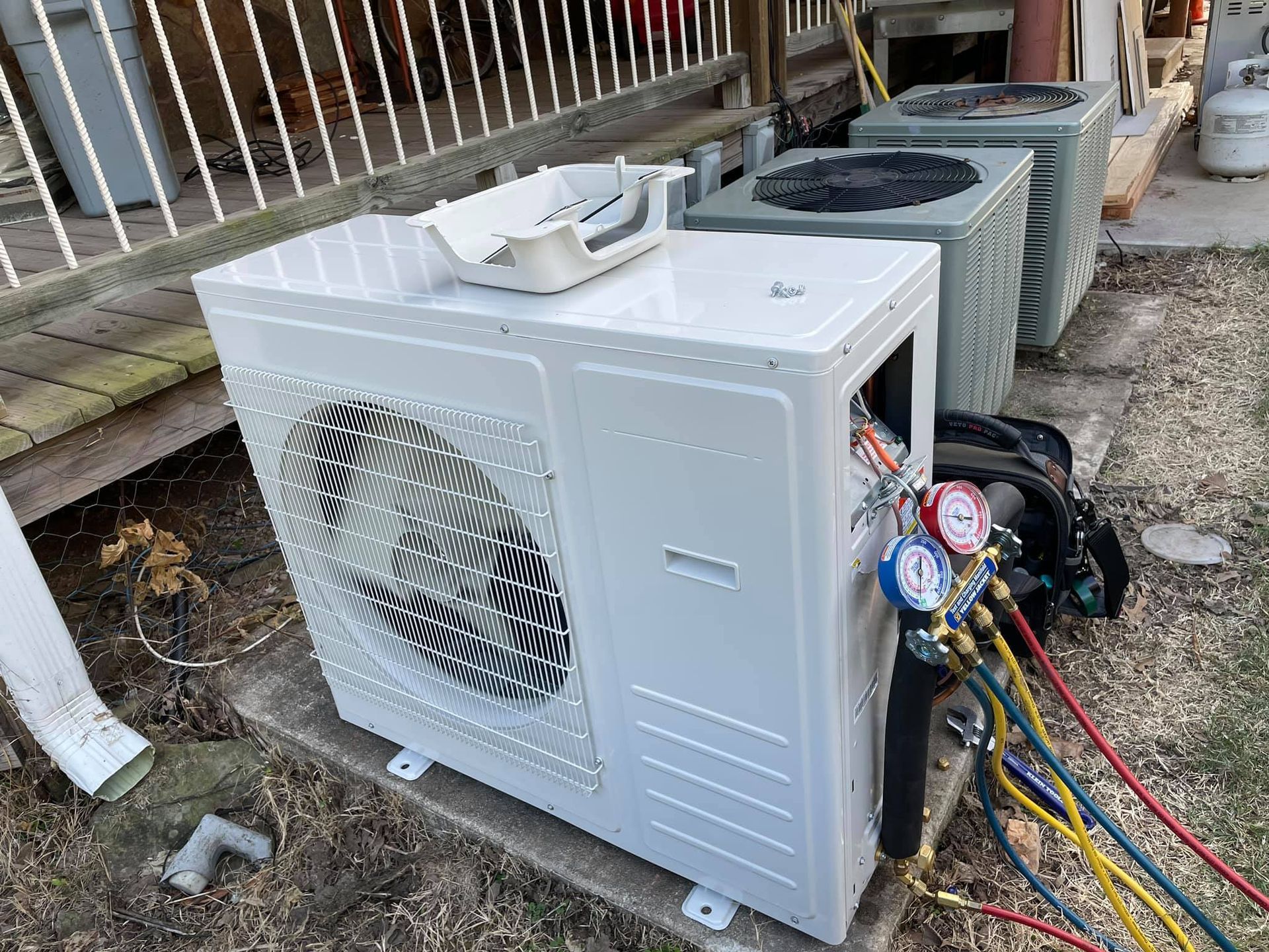 An air conditioning unit being serviced outdoors, with gauges attached to its side. Another unit is in the background.