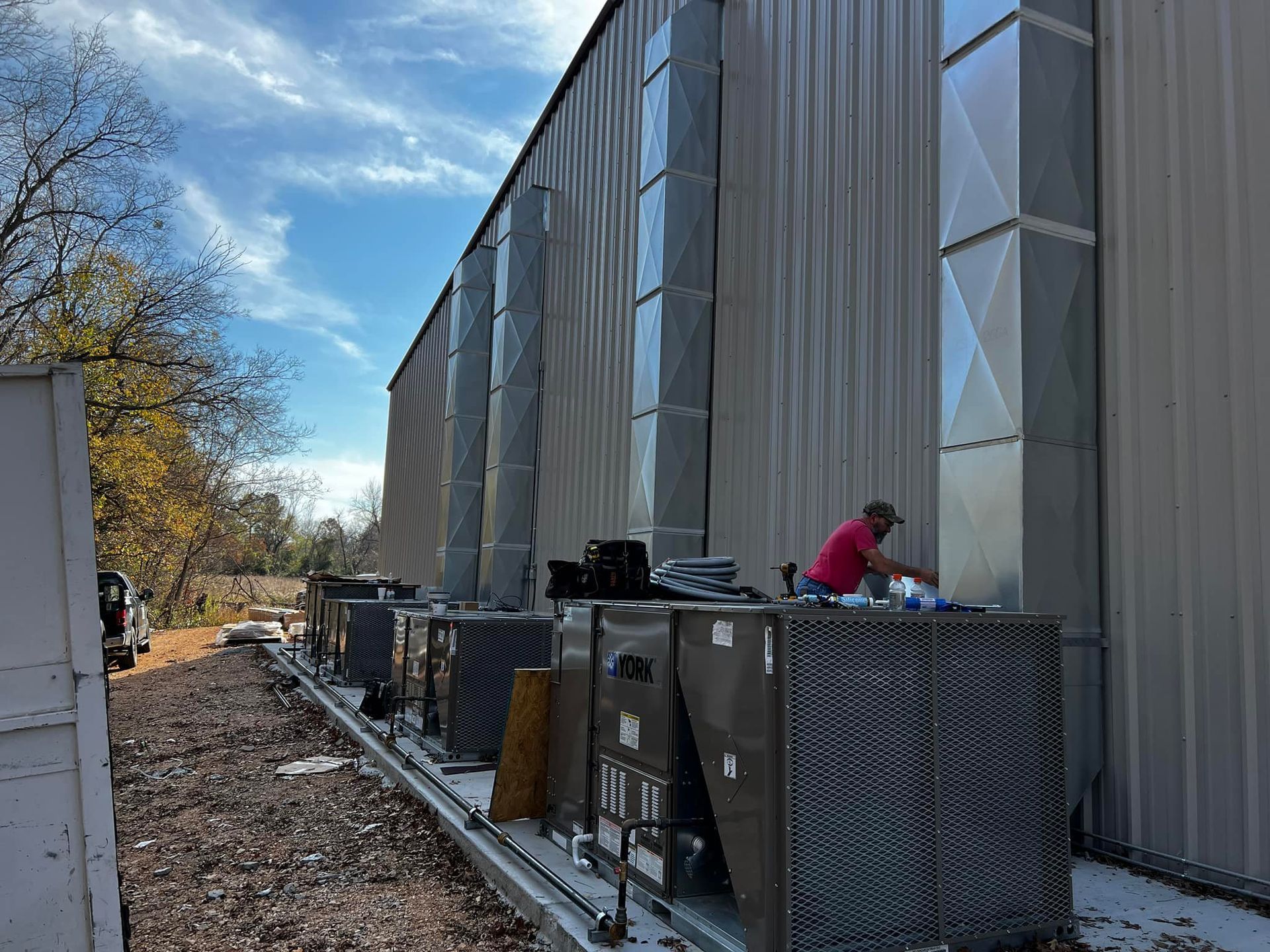 HVAC units installed on the side of a building. A worker in a red shirt is working on the units. Blue sky with some clouds.