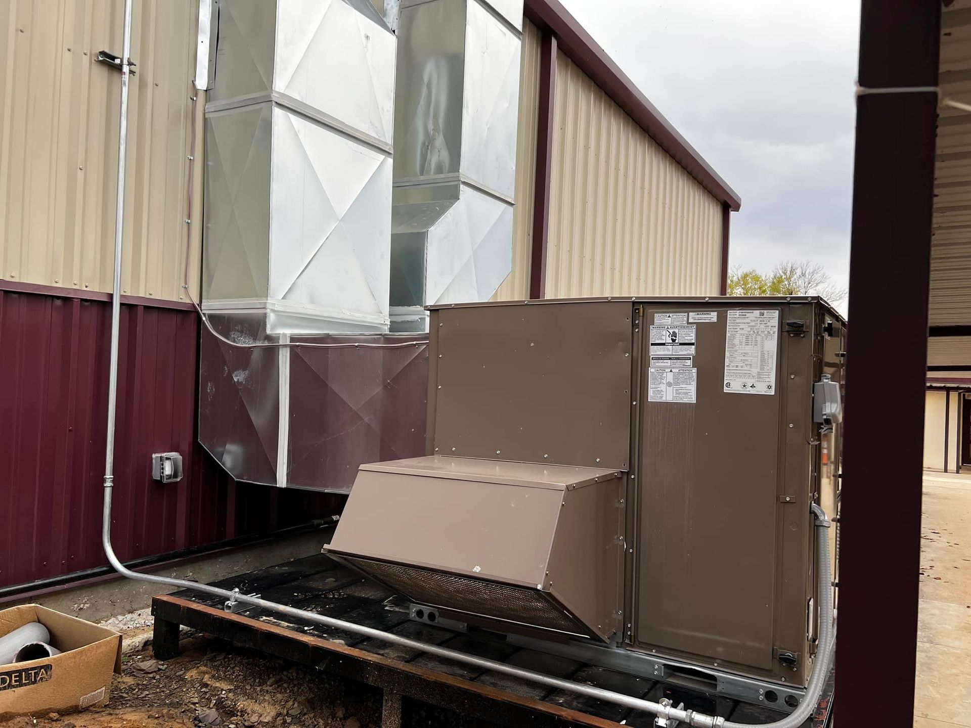 HVAC unit mounted on a metal platform against a building's exterior. Ducts are connected; the building is brown and tan.