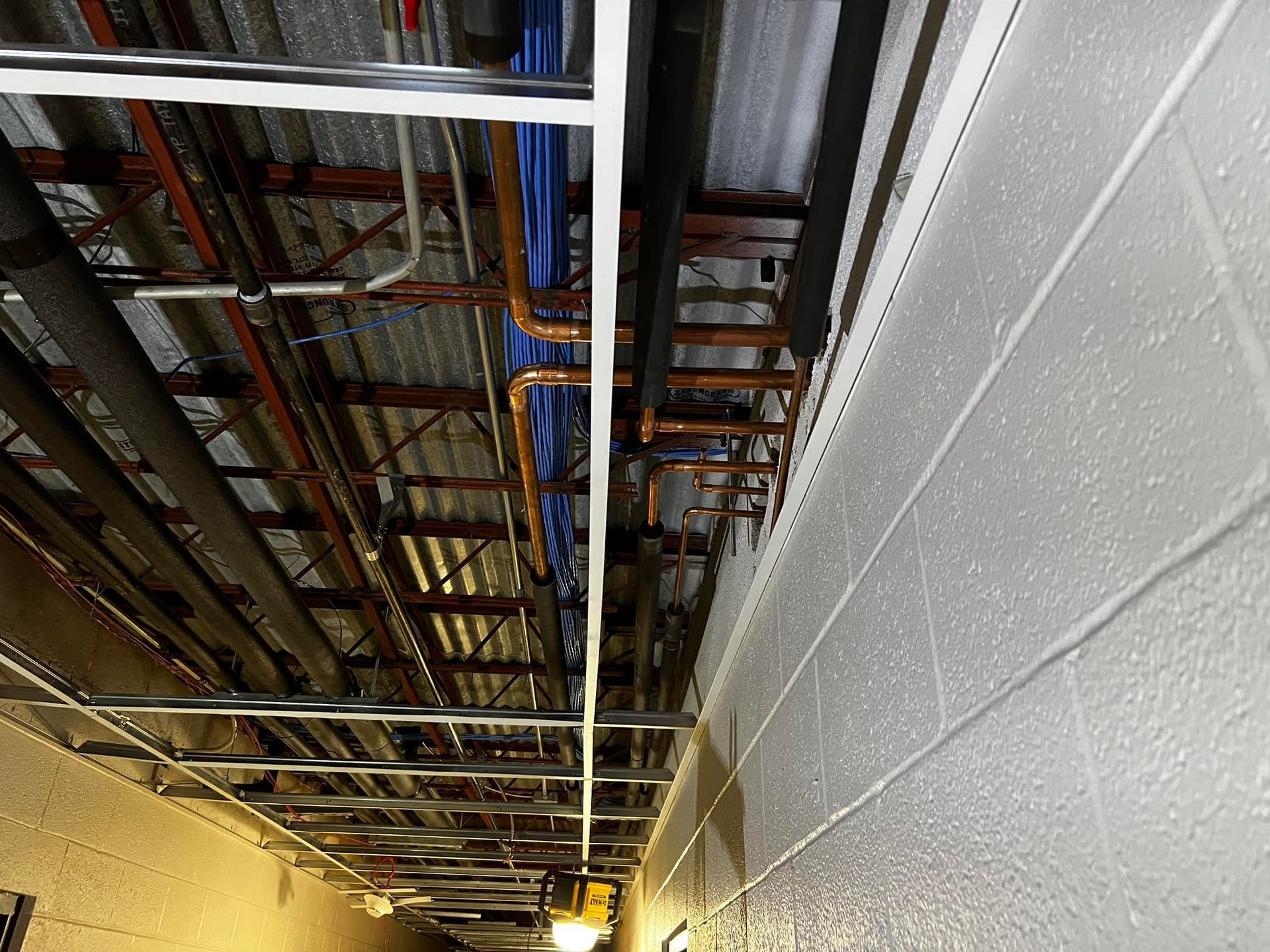 Overhead view of exposed pipes and conduits in a narrow hallway. Copper and black pipes run across a metal ceiling grid.