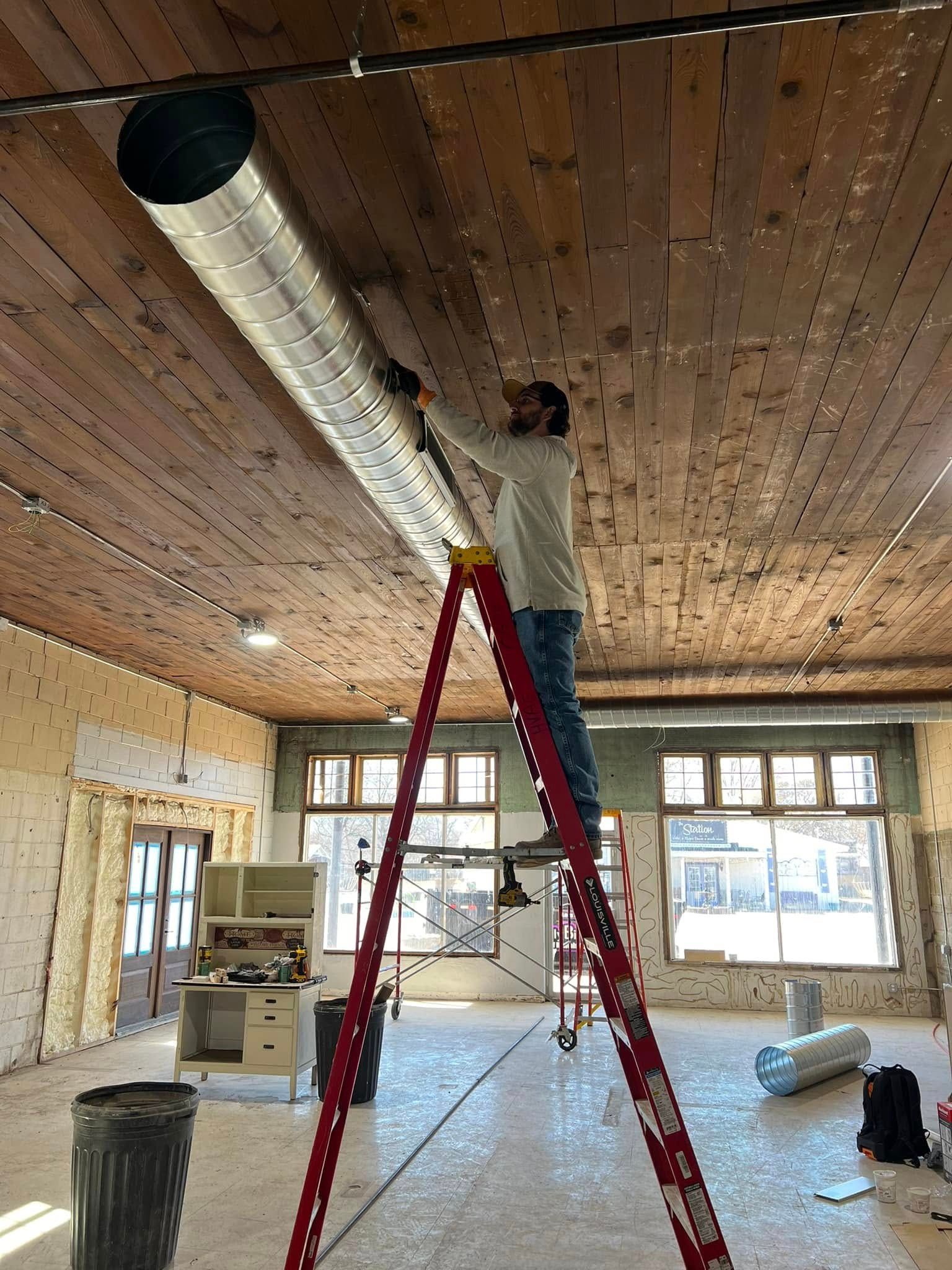 A person installs HVAC ductwork from a ladder in a room with exposed wood ceiling and windows.
