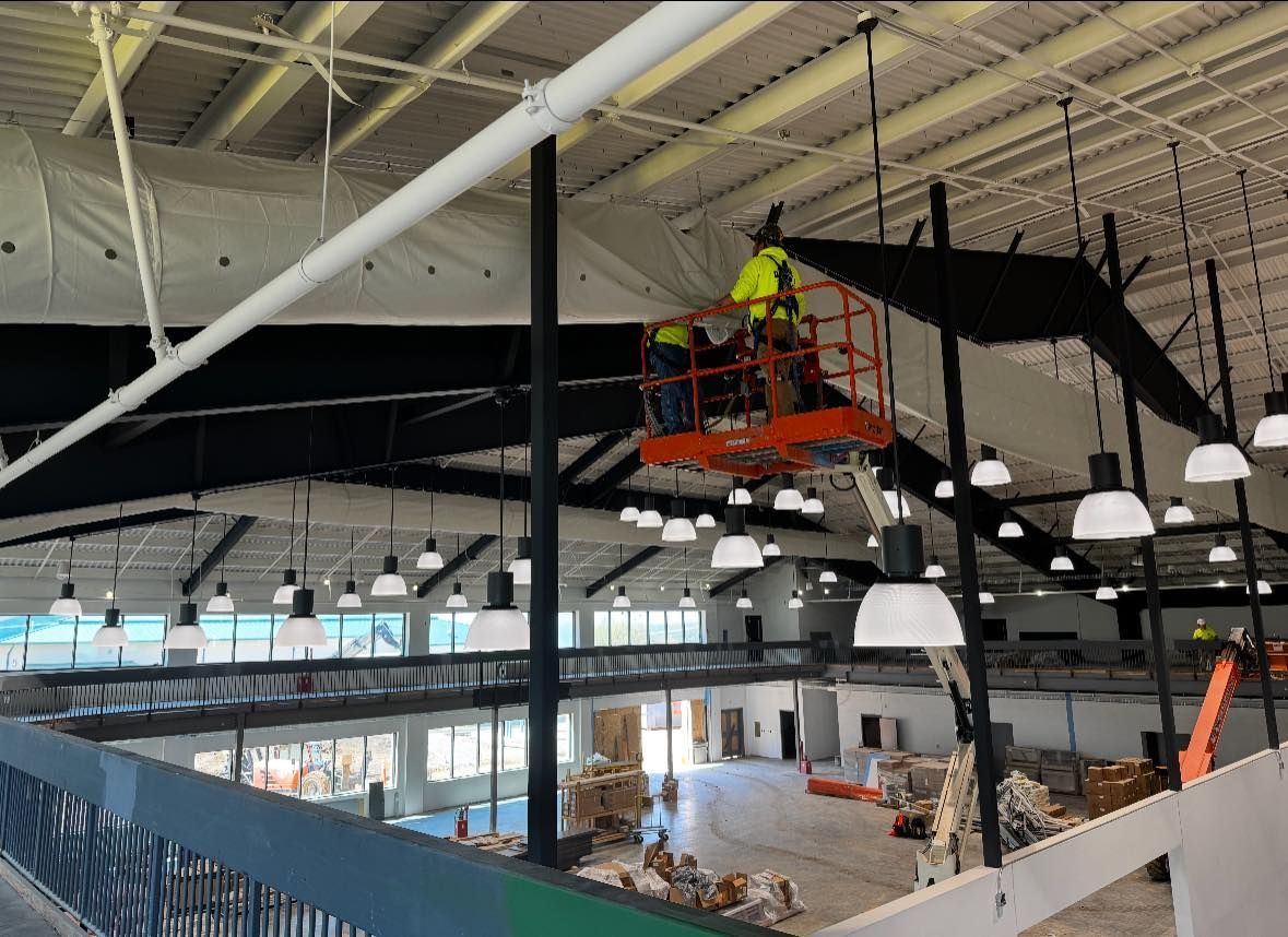 Construction workers in an orange lift working on the ceiling of a large indoor space with exposed beams and hanging lights.