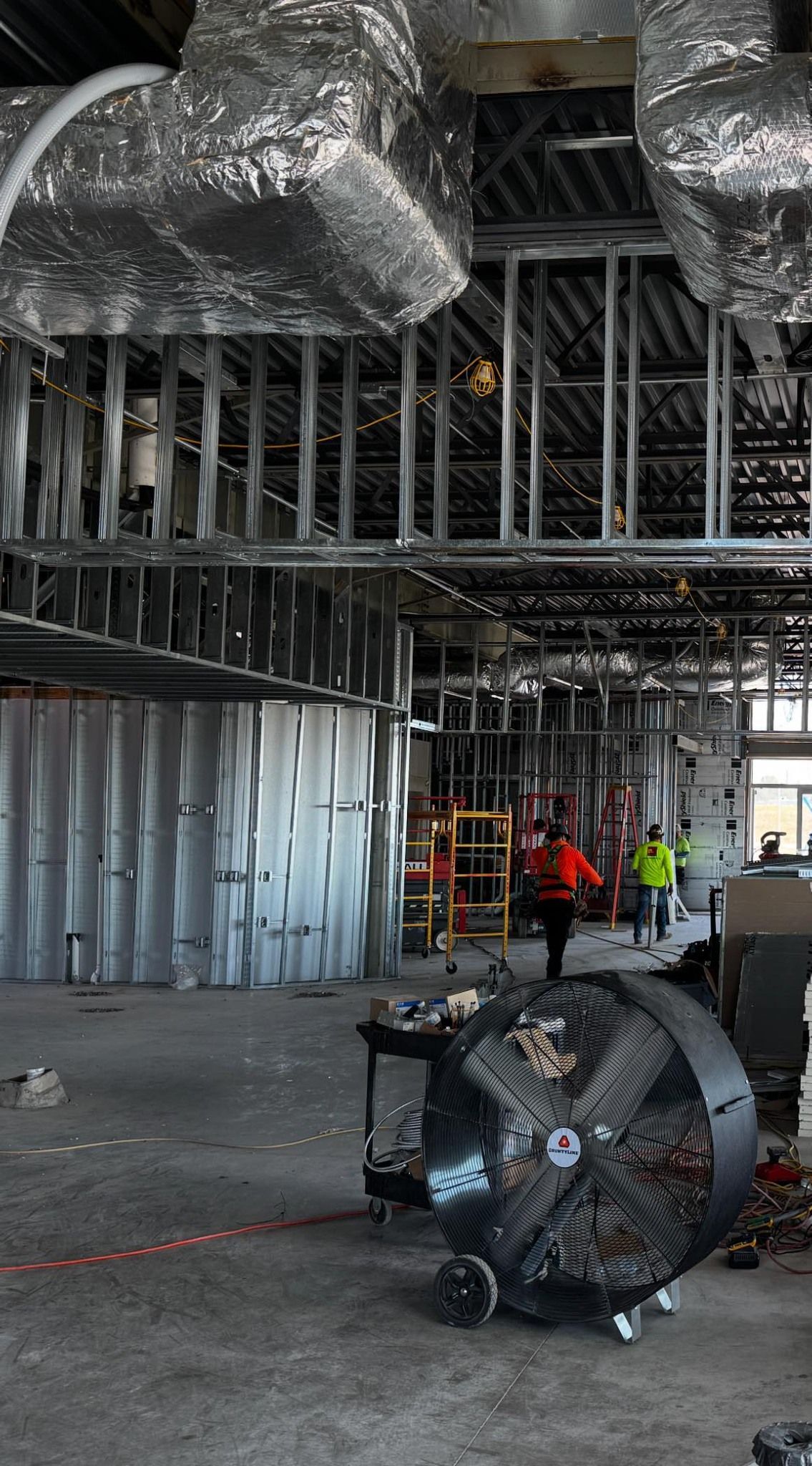 Interior of a building under construction, metal framework visible. Workers in safety vests walk amongst equipment.