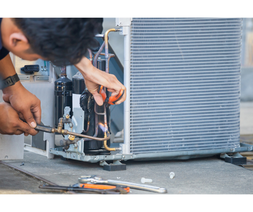 HVAC technician repairing an air conditioning unit with a torch and tools outside, near a metal radiator.