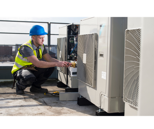A technician in red overalls repairs an air conditioning unit mounted on a white building, using a ladder.