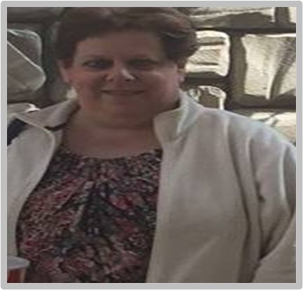 A woman wearing a white jacket and a floral shirt is standing in front of a stone wall.