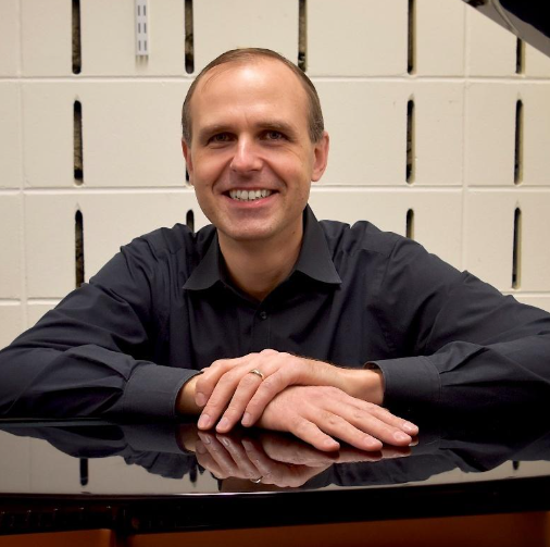 A man is sitting at a desk with his hands folded and smiling