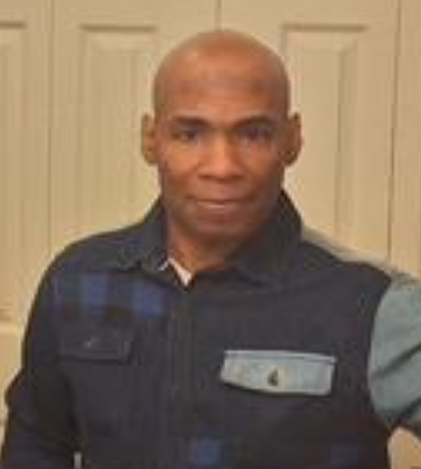 A bald man in a black shirt is standing in front of a white door.