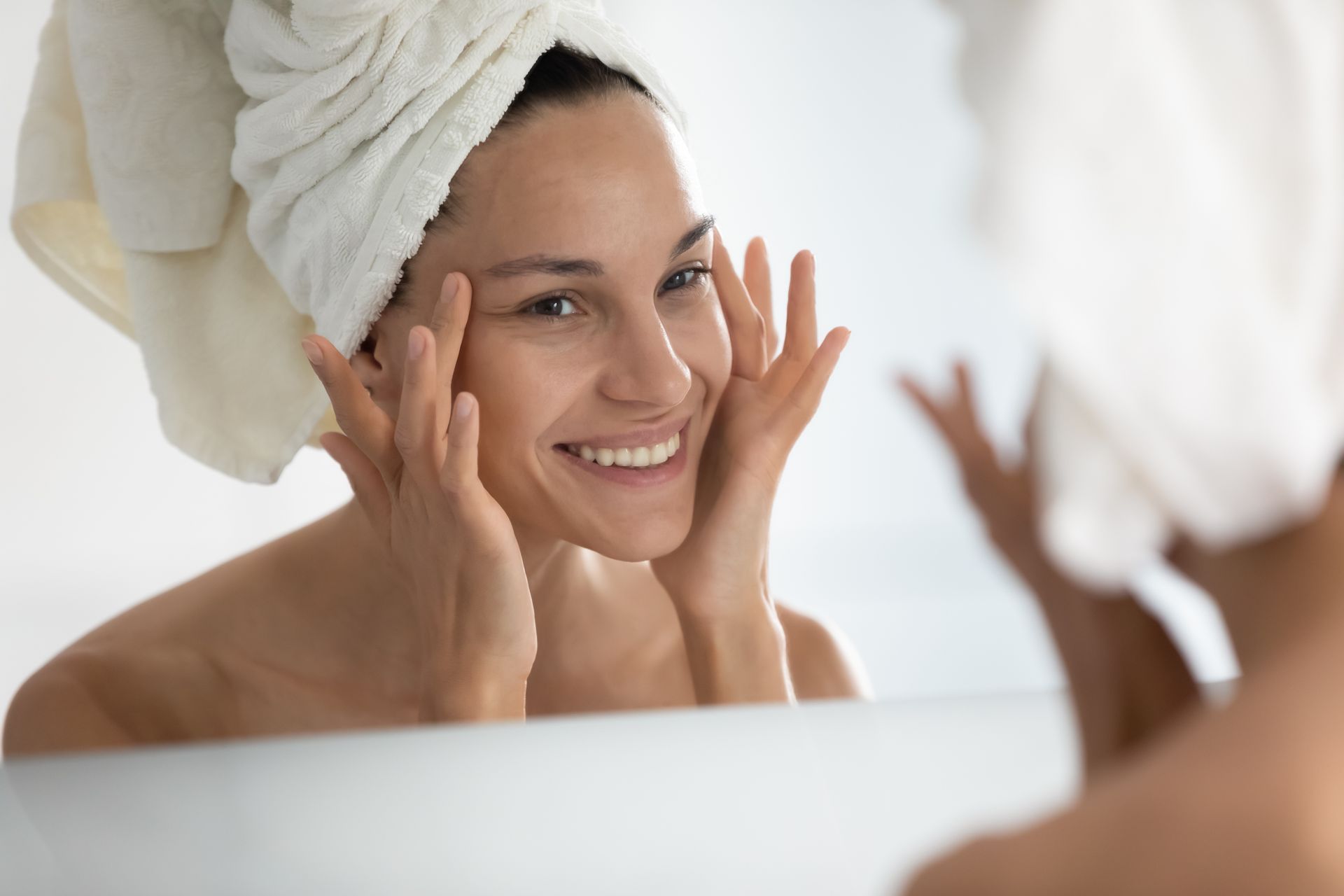 A woman with a towel wrapped around her head is looking at her face in the mirror.