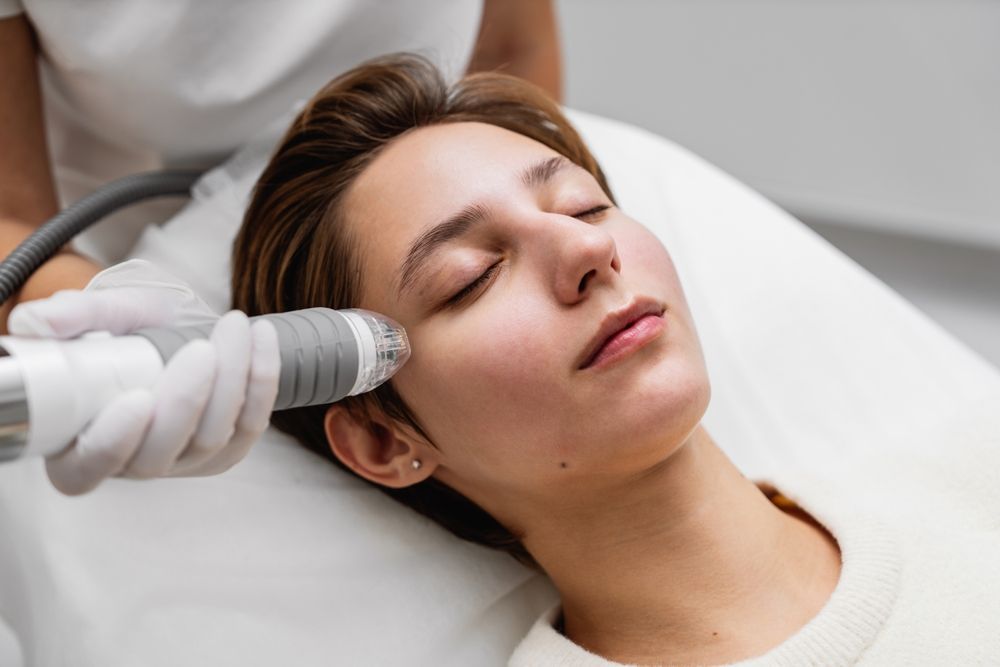 Woman receiving facial treatment in a clinic; a medical device is held near her face.
