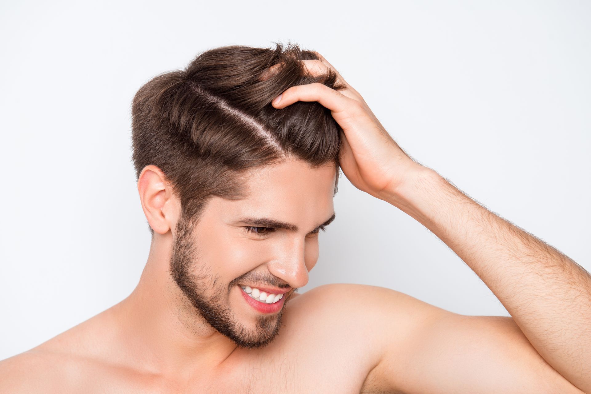 A shirtless man with a beard is smiling and touching his hair.