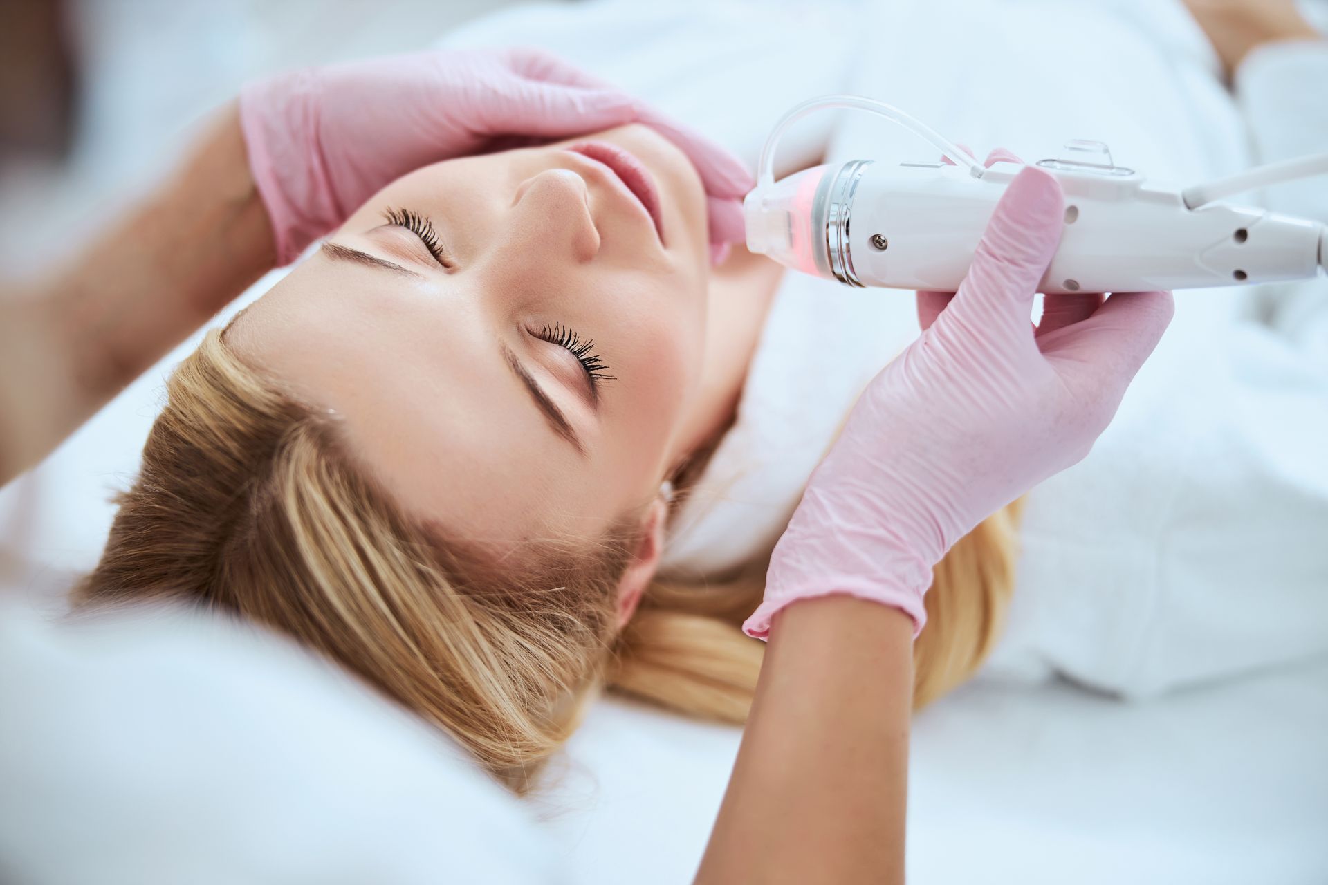 Woman receiving facial treatment with a device; hands in pink gloves, eyes closed, spa setting.