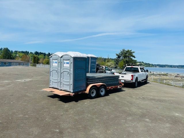 A white truck is towing a trailer with a portable toilet on it.