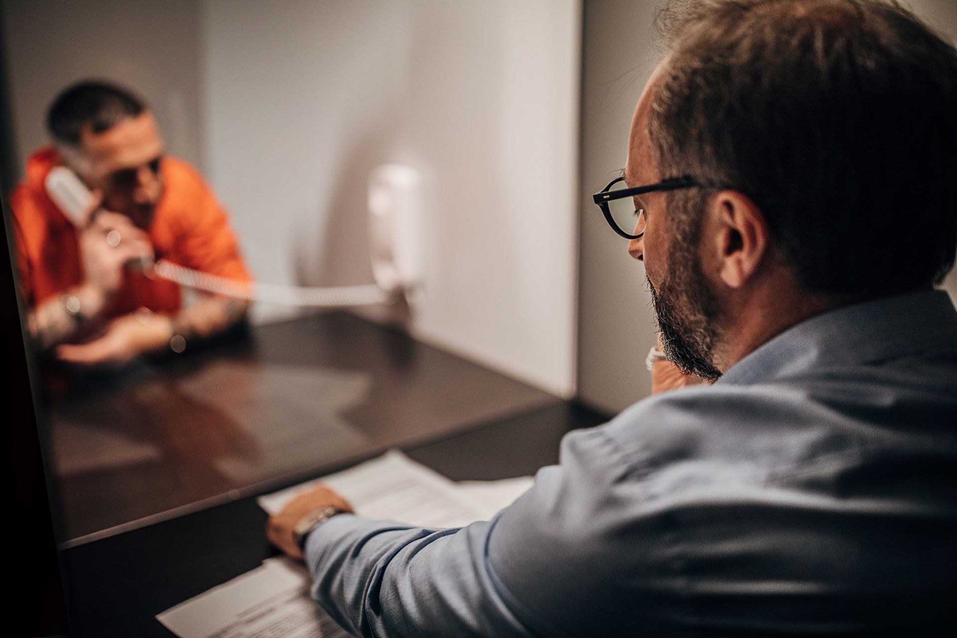 A man is sitting at a desk in front of a mirror talking to a man in an orange shirt.