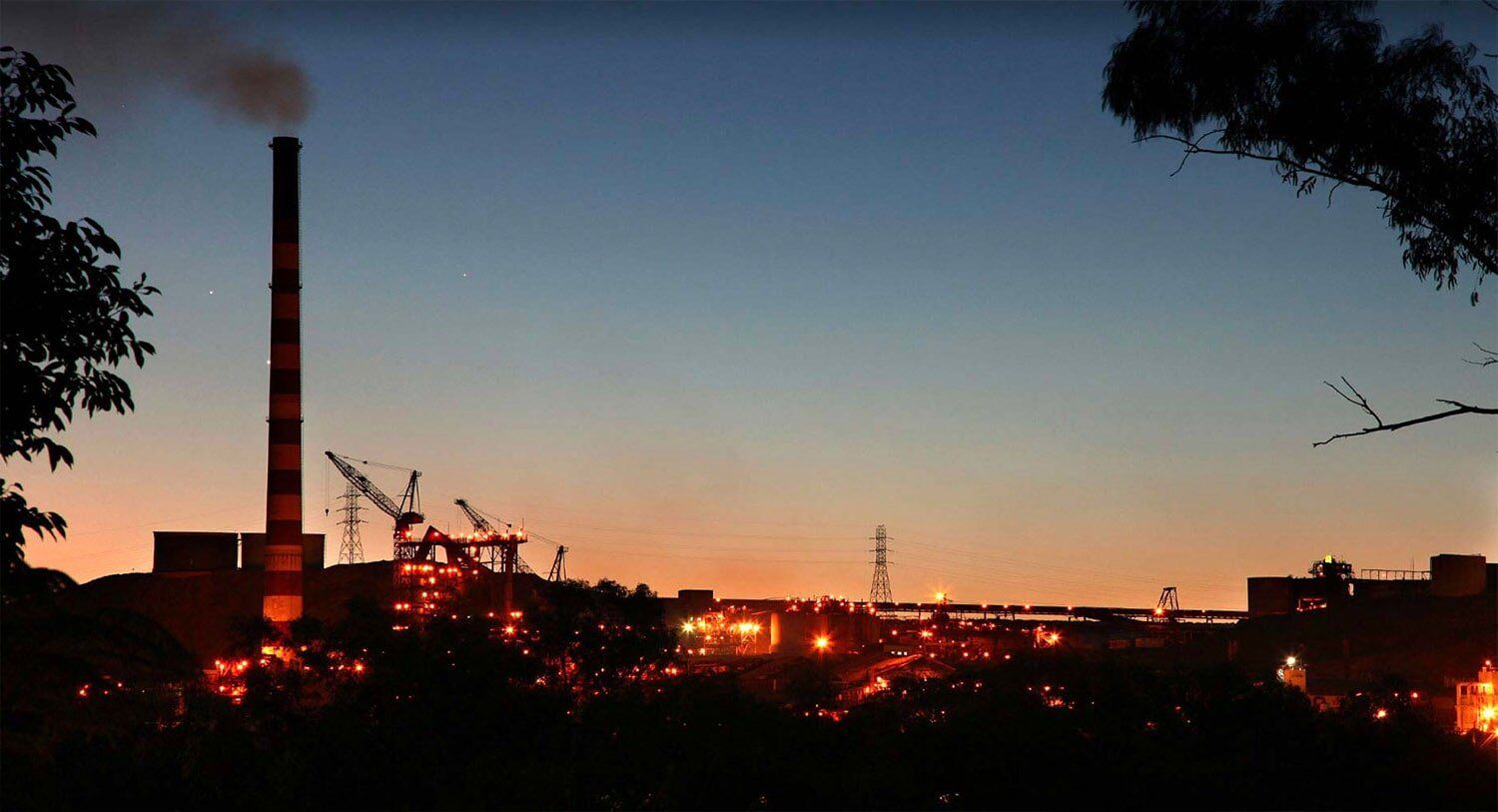 Factory with smokestack emitting smoke at dusk; cranes, structures, and lights silhouetted against a blue and orange sky.