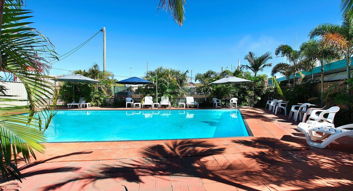 Pool with lounge chairs and umbrellas on a sunny day. Palm tree shadows on the brick.