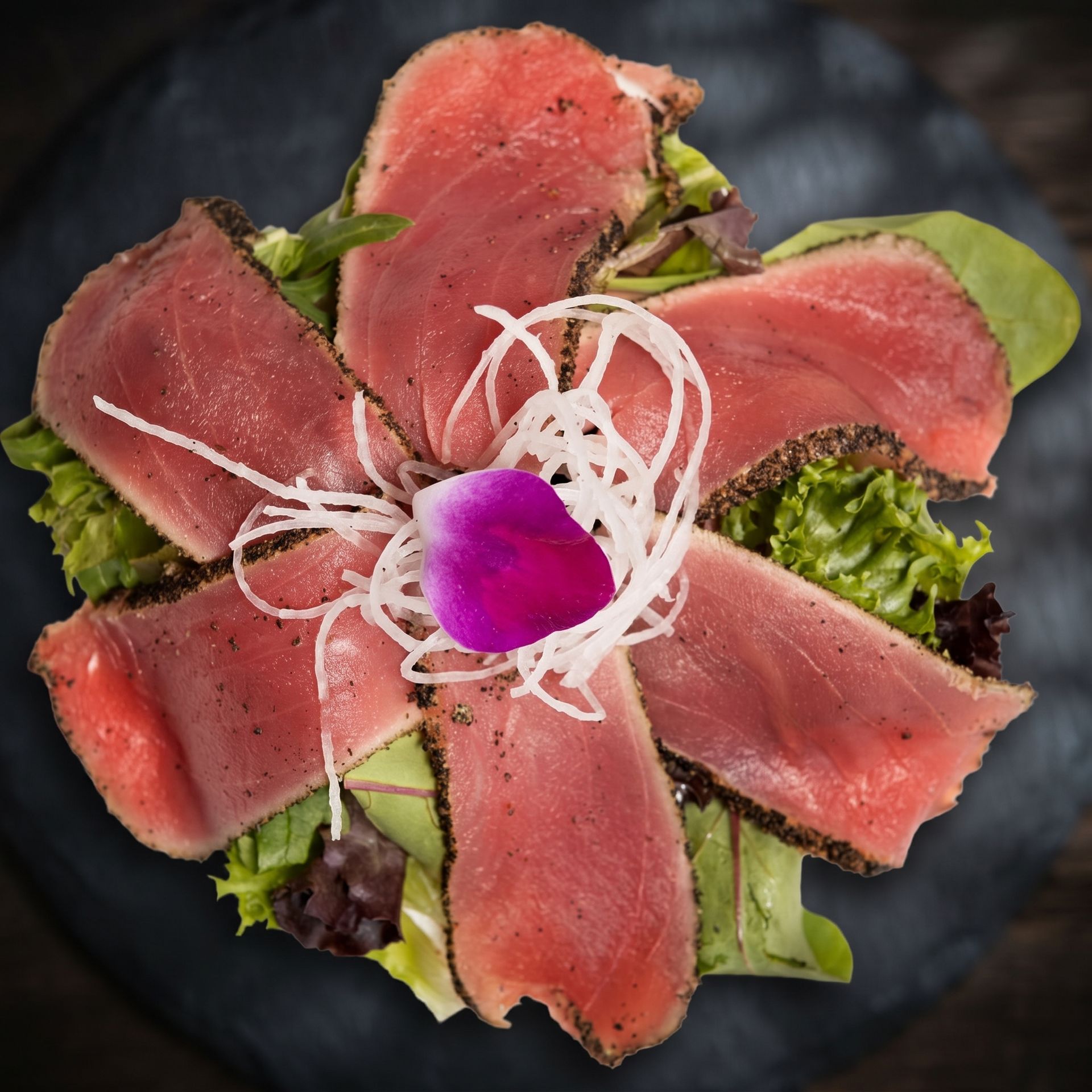 Seared tuna slices arranged around a purple flower on greens, topped with white onion strands