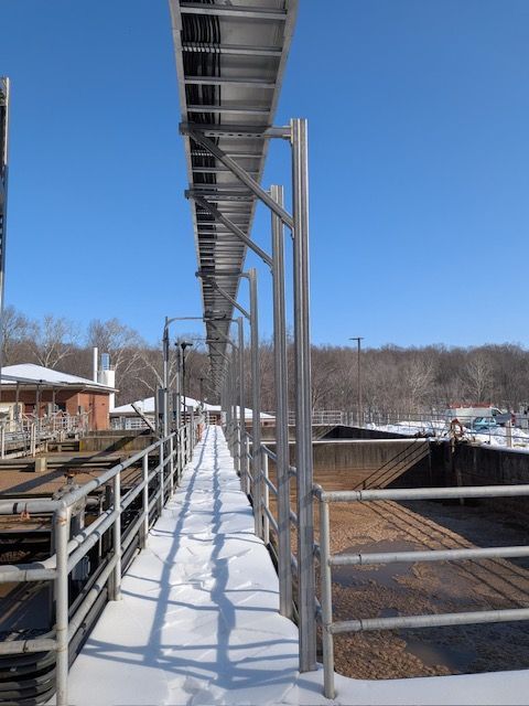 A metallic catwalk with railings covered in snow, leading toward a bright blue sky above an industrial wastewater plant.