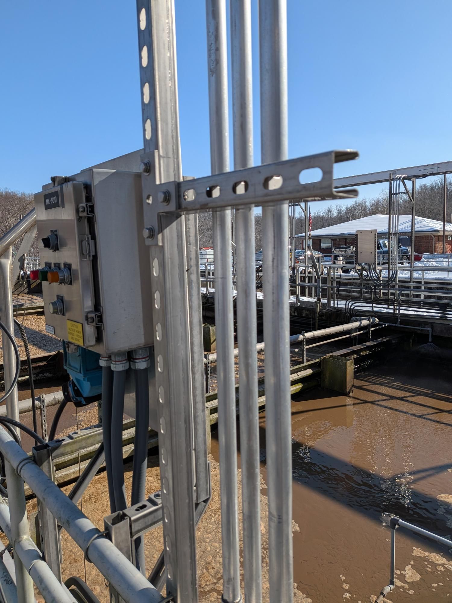 A stainless steel control box and vertical conduits mounted on metal struts overlooking an industrial water basin.