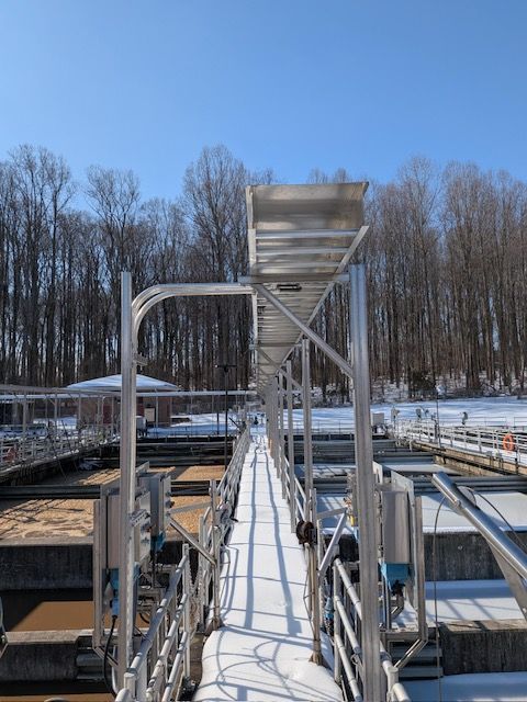 A snow-covered catwalk and metallic structure spanning above concrete water treatment tanks in a winter landscape.