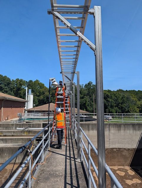 A person wearing a high-visibility vest stands on a ladder, installing electrical conduits above an outdoor walkway.