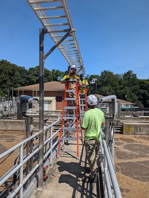 Two technicians work on an overhead cable tray while standing on a ladder on an outdoor industrial walkway.