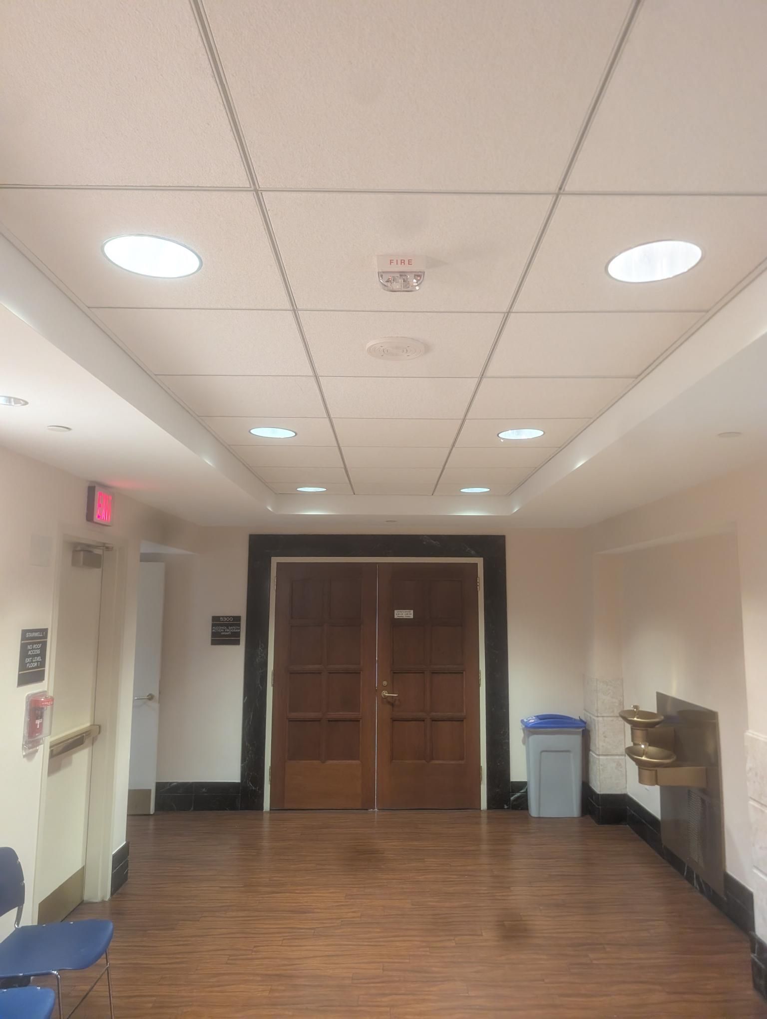 A hallway with light-colored walls and wood-pattern flooring, leading to a set of wooden double doors.