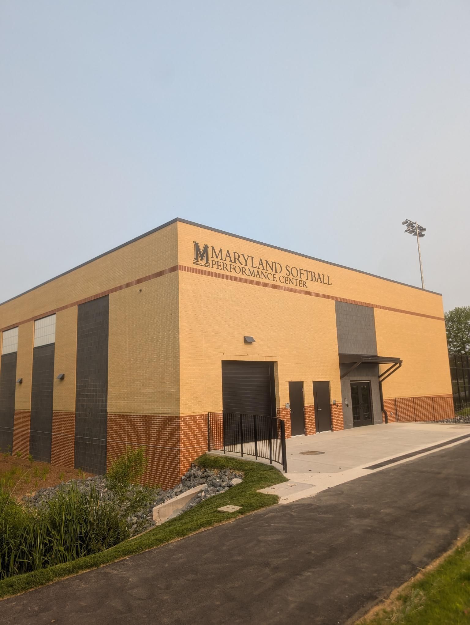 Tan brick Maryland Softball performance center building with a black garage door and walkway on a sunny, hazy day.