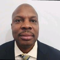 A person with a shaved head and facial hair, wearing a dark suit jacket, light shirt, and striped tie against a white wall.