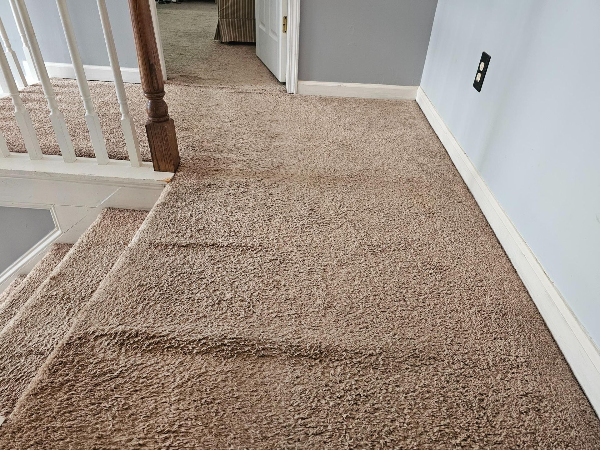 Carpeted stairs and hallway with light brown carpet. A railing is on the left, and a door is visible in the distance.