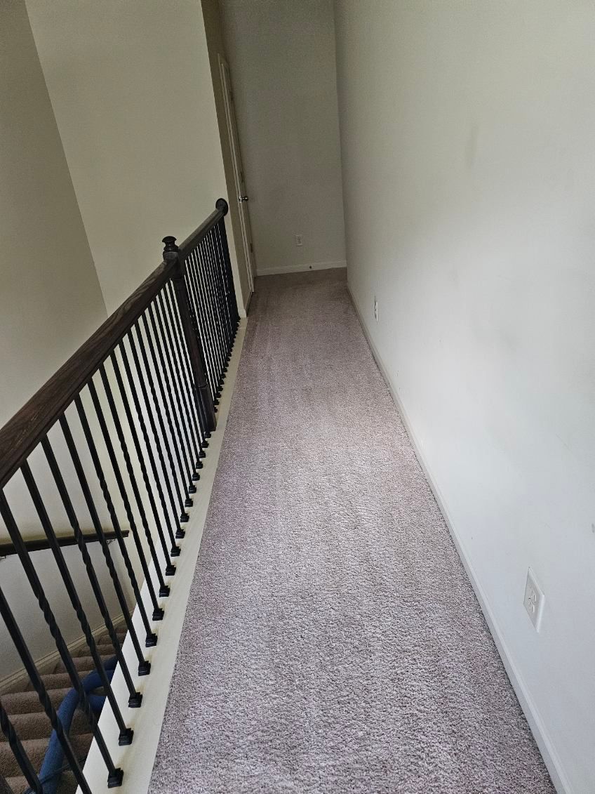 Narrow hallway with carpet, black railing on left, white walls, staircase below.