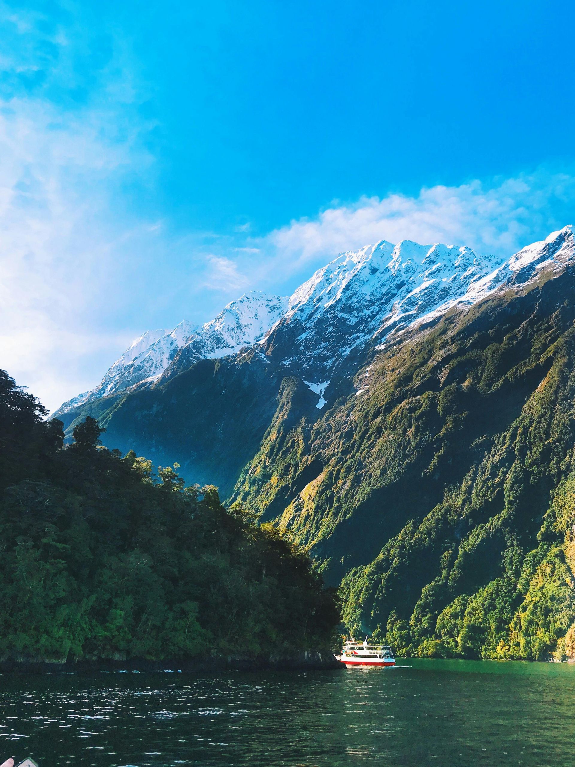 Snow-capped mountain over dark water, green slopes, bright blue sky with a small boat.