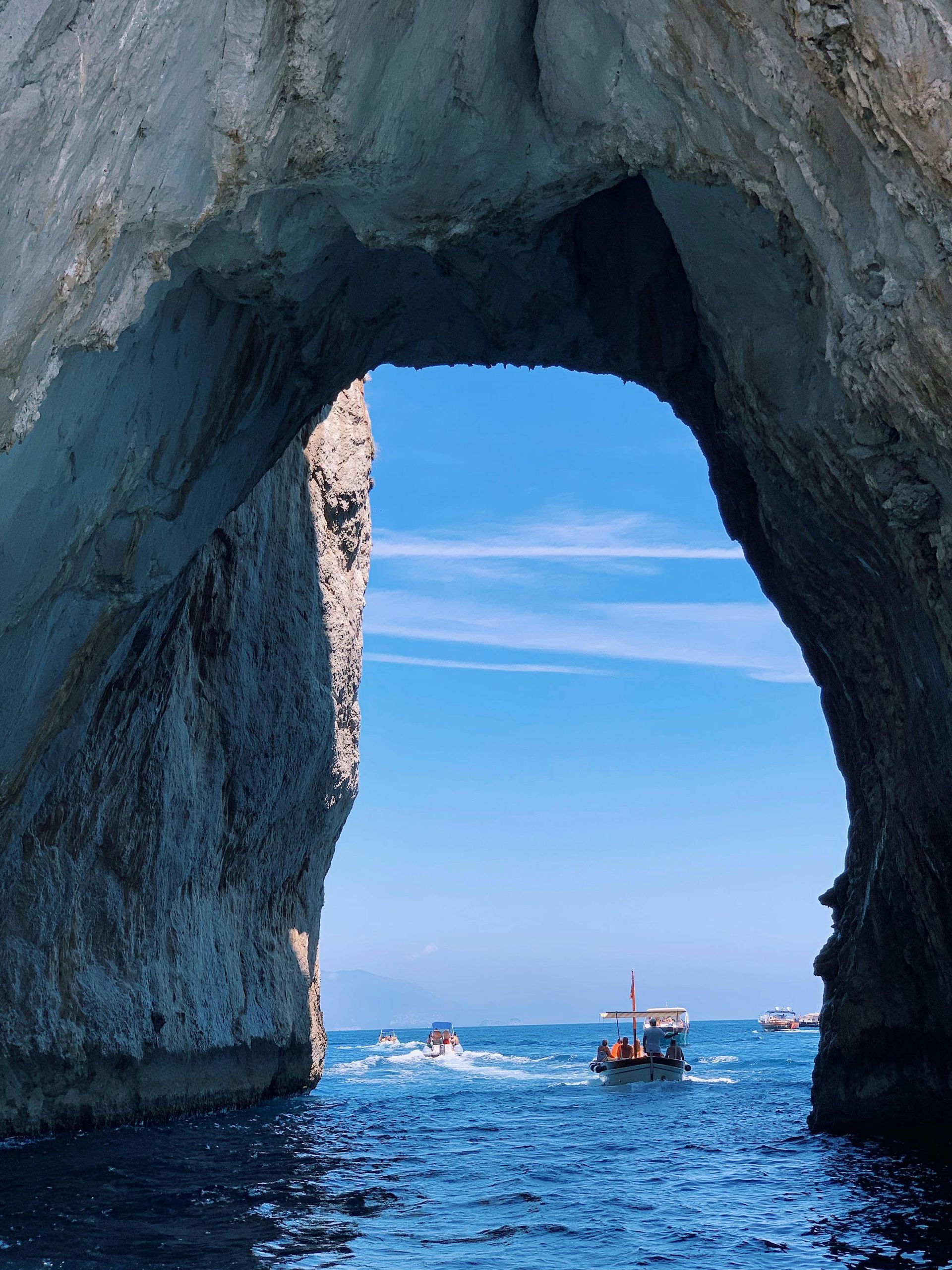 Archway through rock, blue ocean, boats passing, blue sky.