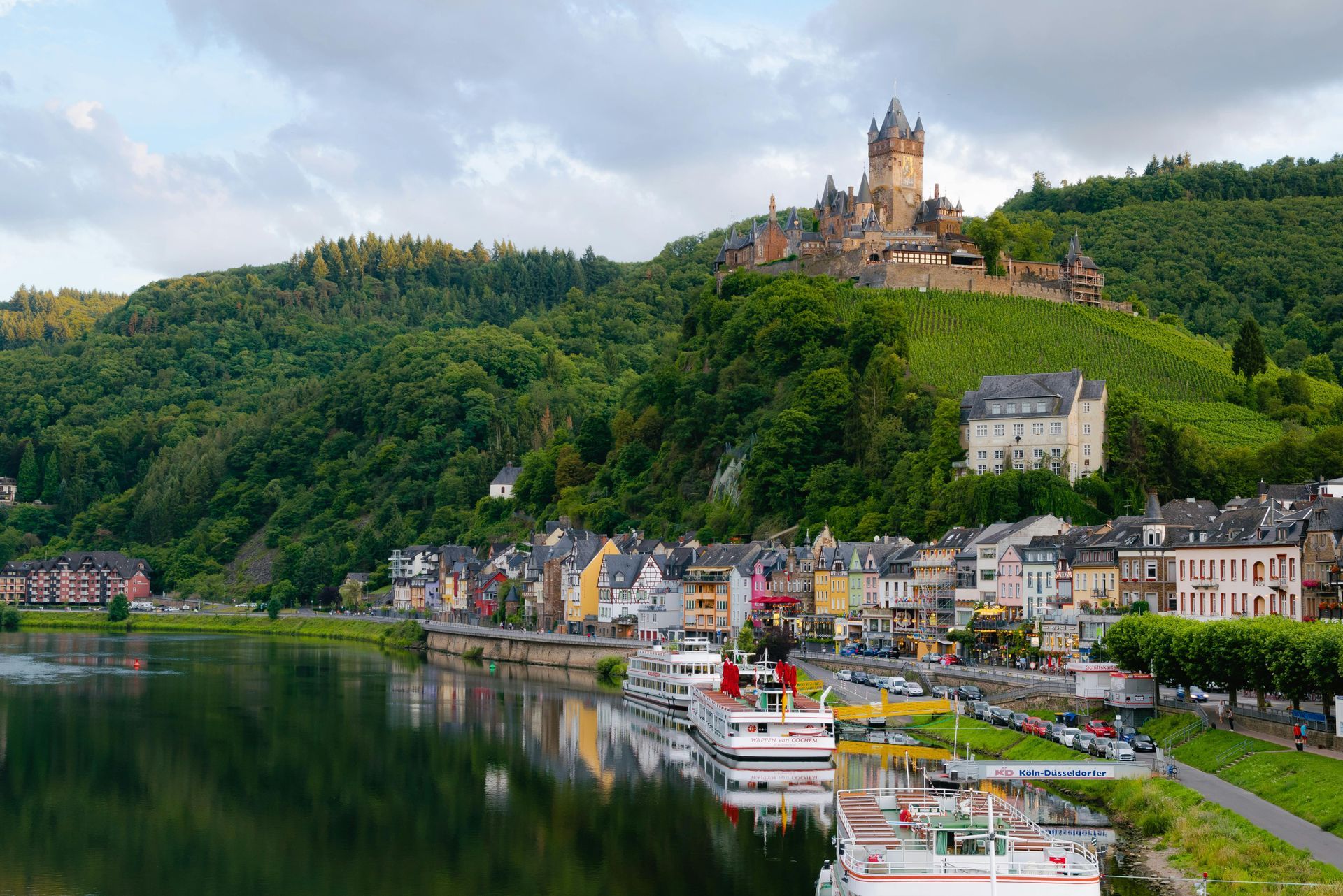 Cochem, Germany. View of the Moselle River, colorful buildings, and Reichsburg Castle atop a green hill under cloudy skies.