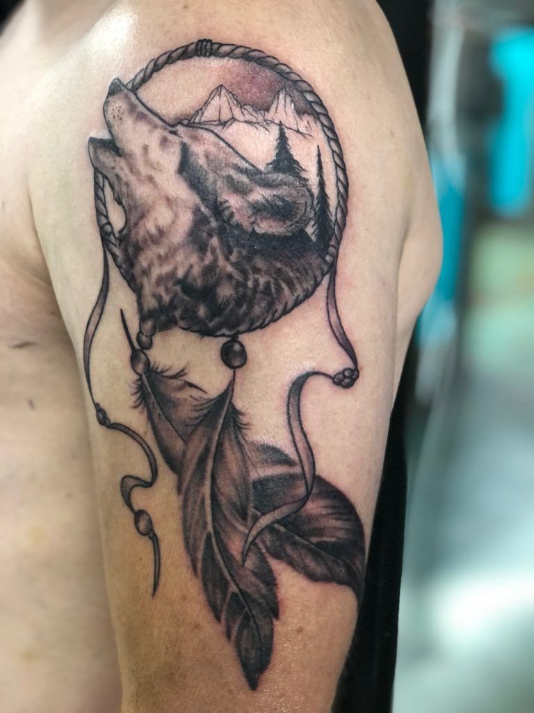 A man has a tattoo of a wolf howling into a dream catcher.