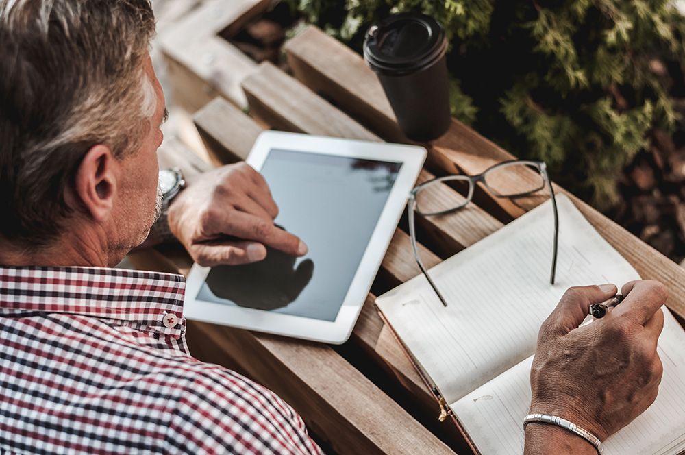 A man is sitting on a bench using a tablet and writing in a notebook.